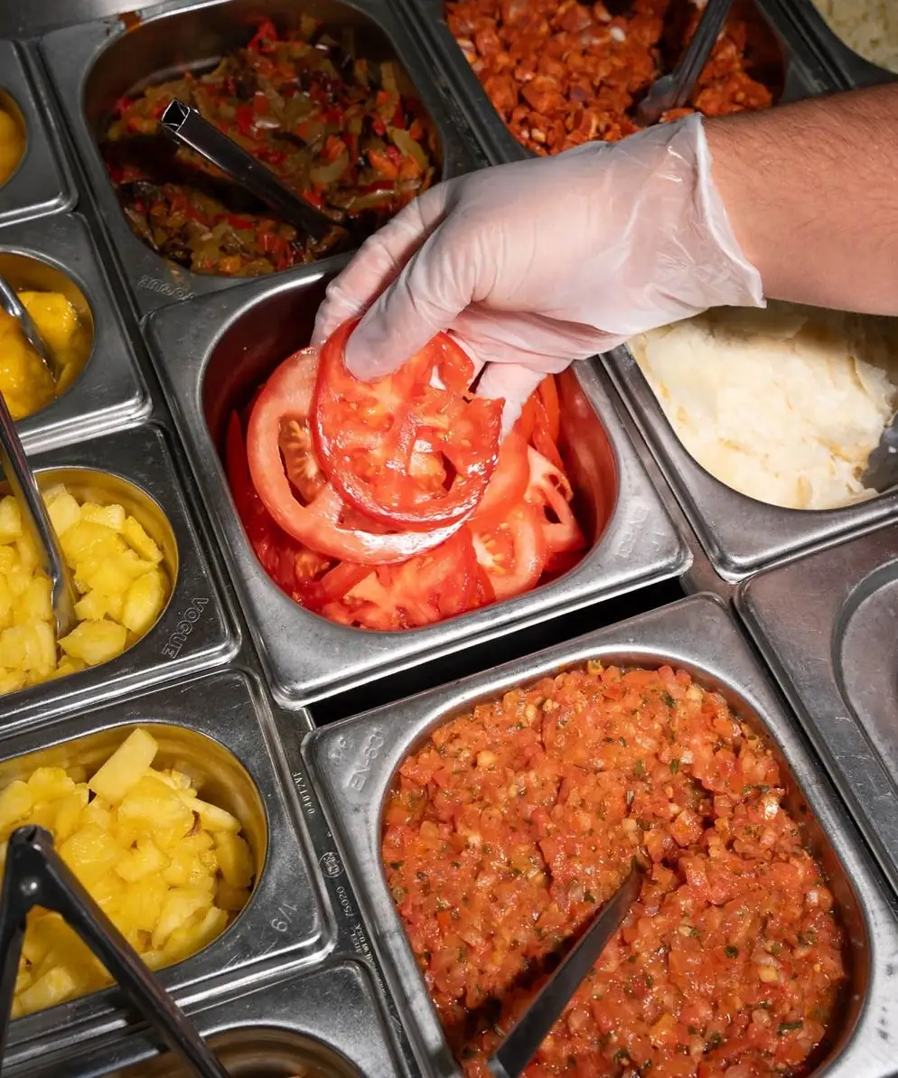 Une main gantée dépose des tranches de tomate dans les bacs du comptoir